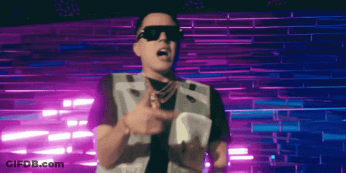 A Man Wearing Sunglasses And A Vest Stands In Front Of A Purple Wall GIF