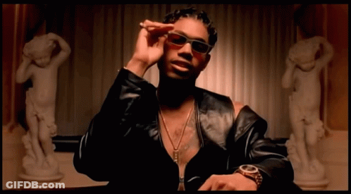 A Man Wearing Sunglasses And A Watch Is Sitting In Front Of A Statue Of A Woman GIF