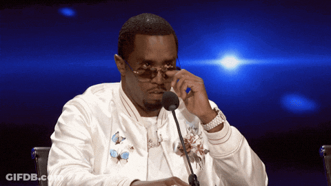 A Man Wearing Sunglasses And A White Jacket Is Sitting At A Table With A Microphone GIF