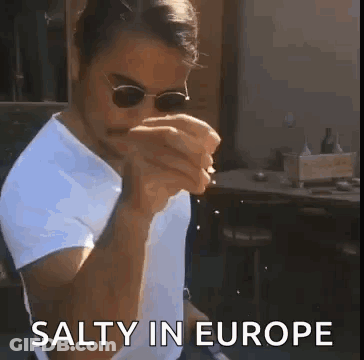A Man Wearing Sunglasses And A White Shirt Is Eating A Sandwich While Sitting At A Table . GIF