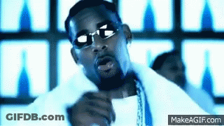 A Man Wearing Sunglasses And A White Shirt Is Singing Into A Microphone In Front Of A Blue Background . GIF