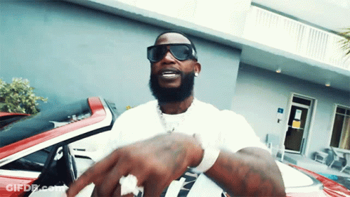 A Man Wearing Sunglasses And A White Shirt Is Standing In Front Of A Red Car GIF