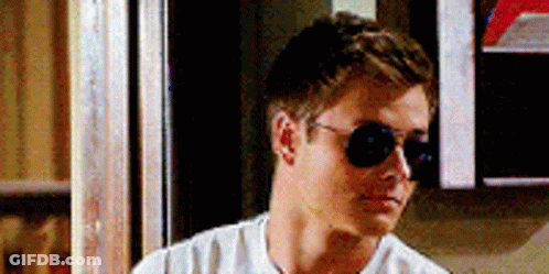 A Man Wearing Sunglasses And A White Shirt Is Standing In Front Of A Sign That Says ' A ' On It . GIF