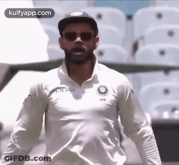A Man Wearing Sunglasses And A White Shirt Is Standing In Front Of A Stadium . GIF