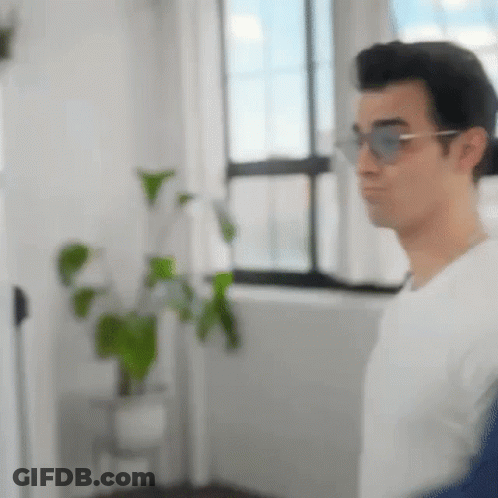 A Man Wearing Sunglasses And A White Shirt Is Standing In Front Of A Window In A Room . GIF