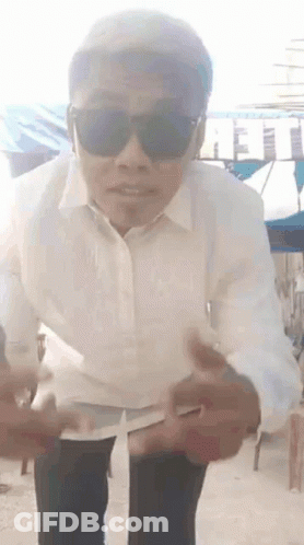 A Man Wearing Sunglasses And A White Shirt Is Standing On A Sidewalk . GIF