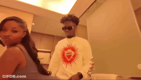 A Man Wearing Sunglasses And A White Shirt With A Red Heart On It Stands Next To A Woman GIF
