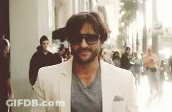A Man Wearing Sunglasses And A White Suit Is Walking Down A Street . GIF