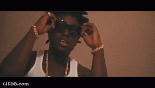 A Man Wearing Sunglasses And A White Tank Top Has Gold Chains Around His Neck GIF