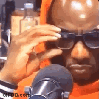 A Man Wearing Sunglasses And An Orange Hoodie Is Sitting In Front Of A Microphone . GIF