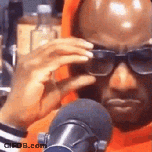 A Man Wearing Sunglasses And An Orange Hoodie Is Sitting In Front Of A Microphone . GIF