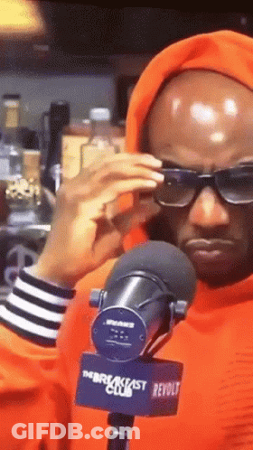 A Man Wearing Sunglasses And An Orange Hoodie Stands In Front Of A Microphone That Says The Breakfast Club GIF