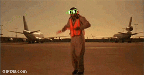 A Man Wearing Sunglasses And An Orange Vest Stands In Front Of Planes GIF