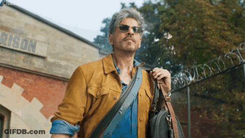 A Man Wearing Sunglasses And Carrying A Briefcase Walks In Front Of A Building That Says Prison GIF
