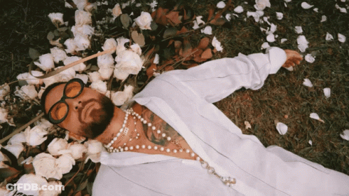 A Man Wearing Sunglasses And Pearls Is Laying In The Grass Surrounded By Flowers GIF