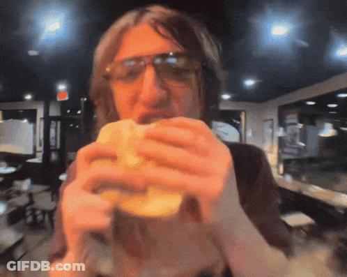 A Man Wearing Sunglasses Eating A Sandwich In A Restaurant GIF