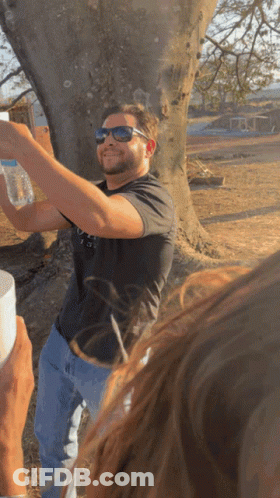 A Man Wearing Sunglasses Holds A Bottle Of Water In Front Of A Tree GIF