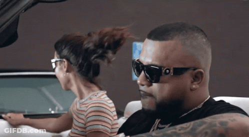 A Man Wearing Sunglasses Is Driving A Car Next To A Woman In A Striped Shirt GIF