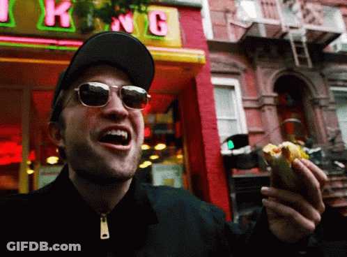 A Man Wearing Sunglasses Is Eating A Hot Dog In Front Of A Store Called King GIF