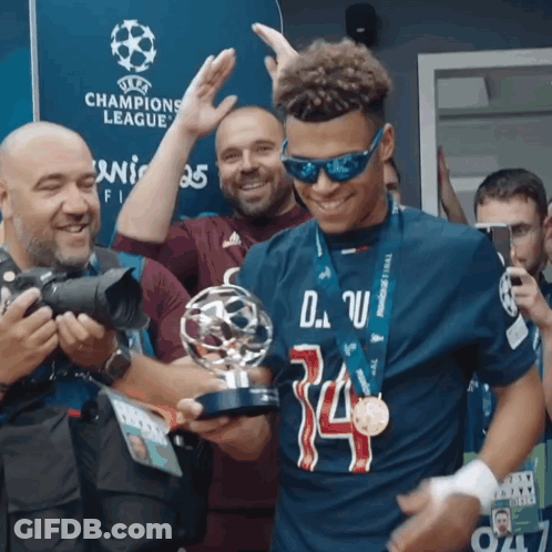 A Man Wearing Sunglasses Is Holding A Trophy In Front Of A Sign That Says Champions League GIF