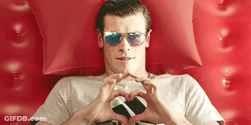 A Man Wearing Sunglasses Is Making A Heart Shape With His Hands While Laying On A Bed . GIF