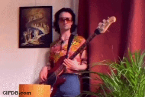 A Man Wearing Sunglasses Is Playing A Bass Guitar In A Living Room . GIF