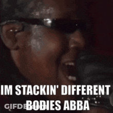 A Man Wearing Sunglasses Is Singing Into A Microphone And Says I 'M Stackin ' Different Bodies Abba . GIF