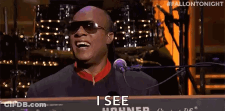 A Man Wearing Sunglasses Is Singing Into A Microphone While Playing A Piano . GIF
