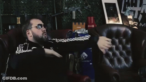 A Man Wearing Sunglasses Is Sitting In A Chair In Front Of A Sign That Says Am GIF