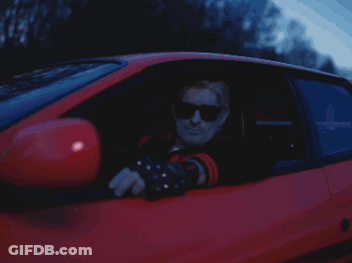 A Man Wearing Sunglasses Is Sitting In The Driver 'S Seat Of A Red Car GIF