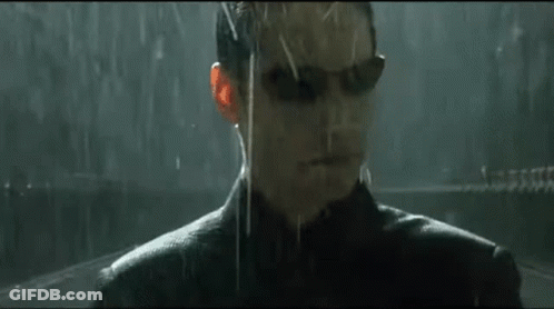 A Man Wearing Sunglasses Is Standing In The Rain Looking At The Camera . GIF