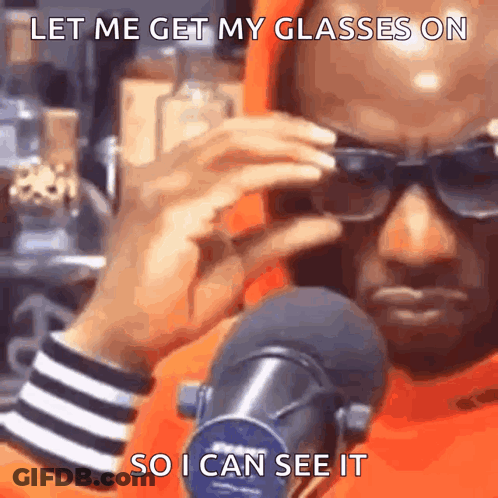 A Man Wearing Sunglasses Is Talking Into A Microphone While Holding His Glasses On . GIF