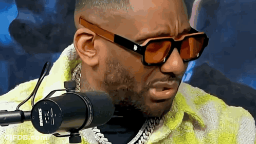 A Man Wearing Sunglasses Is Talking Into A Shure Microphone . GIF