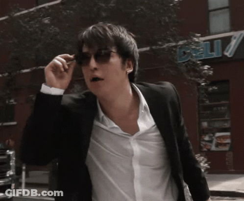A Man Wearing Sunglasses Is Walking Down The Street In Front Of A Deli GIF