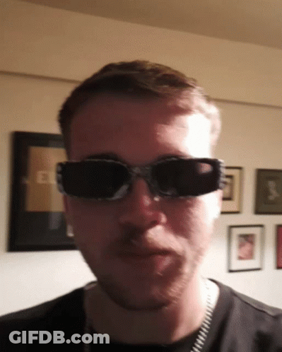 A Man Wearing Sunglasses Looks At The Camera In Front Of A Wall With Pictures On It GIF