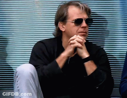 A Man Wearing Sunglasses Sits With His Hands Folded In Front Of A Window . GIF