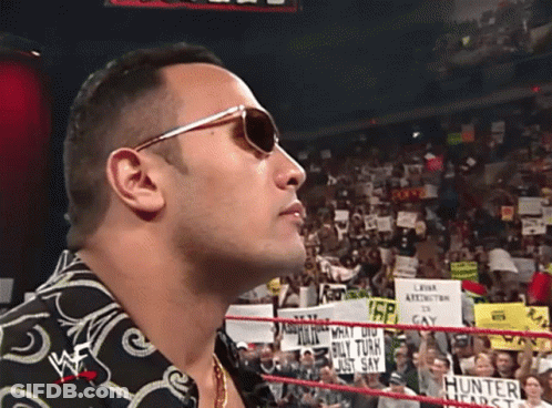 A Man Wearing Sunglasses Stands In A Wrestling Ring With A Crowd Behind Him GIF