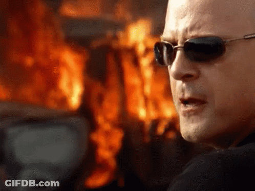 A Man Wearing Sunglasses Stands In Front Of A Burning Building GIF