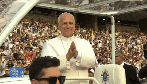 A Man Wearing Sunglasses Stands In Front Of A Crowd Of People While The Pope Applauds In The Background GIF