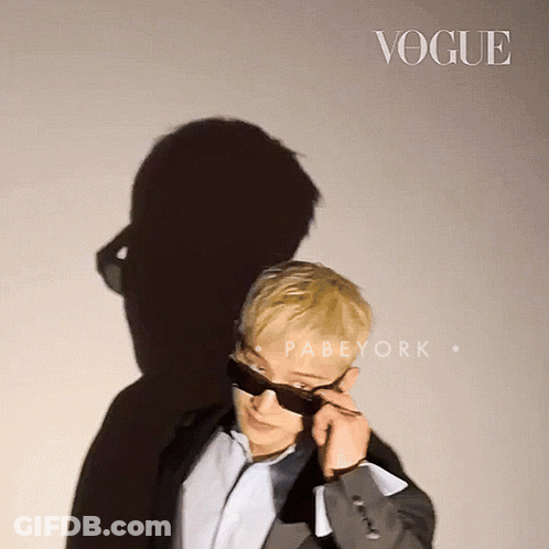 A Man Wearing Sunglasses Stands In Front Of A Wall That Says Vogue GIF