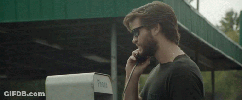 A Man Wearing Sunglasses Talks On A Pay Phone GIF
