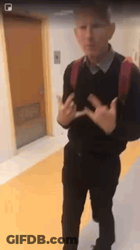 A Man With A Backpack Is Standing In A Hallway Making A Peace Sign With His Hands . GIF