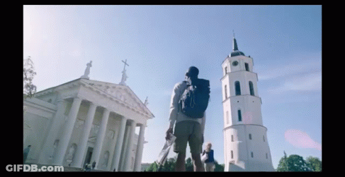 A Man With A Backpack Stands In Front Of A Large Building With Columns And A Clock Tower GIF