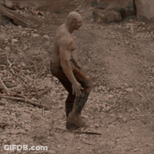 A Man With A Bald Head Is Standing In The Dirt Holding A Sword . GIF
