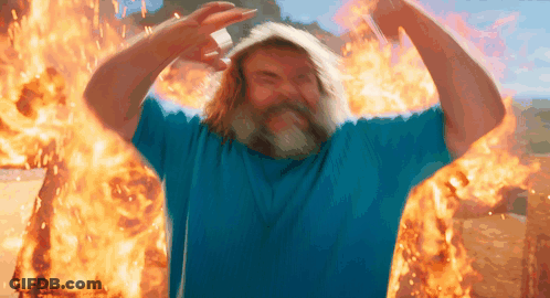 A Man With A Beard And A Blue Shirt Is Standing In Front Of A Fire GIF