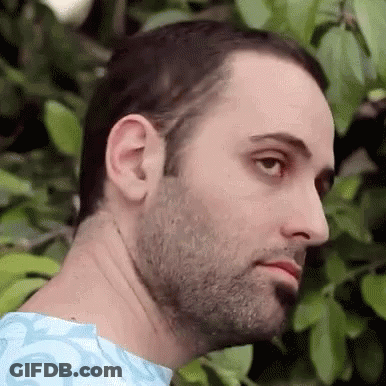 A Man With A Beard And A Blue Shirt Is Standing In Front Of A Tree And Looking At The Camera . GIF