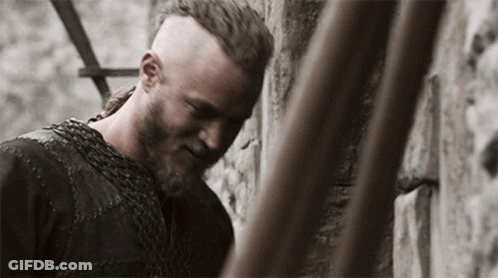 A Man With A Beard And A Chain Around His Neck Is Leaning Against A Wall GIF