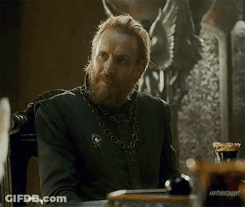 A Man With A Beard And A Chain Around His Neck Is Sitting At A Table In A Chair . GIF