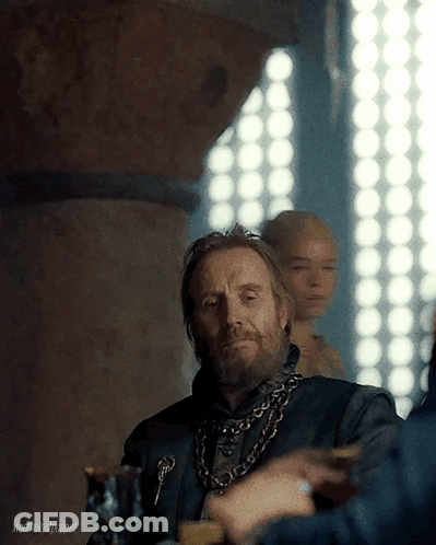 A Man With A Beard And A Chain Around His Neck Is Sitting At A Table With Other People . GIF