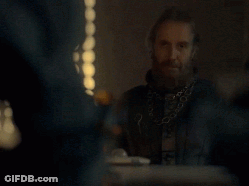 A Man With A Beard And A Chain Around His Neck Is Sitting At A Table With Other People . GIF
